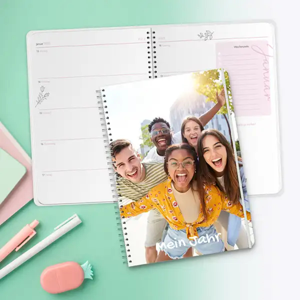 Personalisierter Schülerkalender mit Spiralbindung und individuellem Fotocover 'Mein Jahr', liegend vor einem geöffneten Kalender mit Wochenansicht auf mintgrünem Hintergrund mit Schreibwaren.